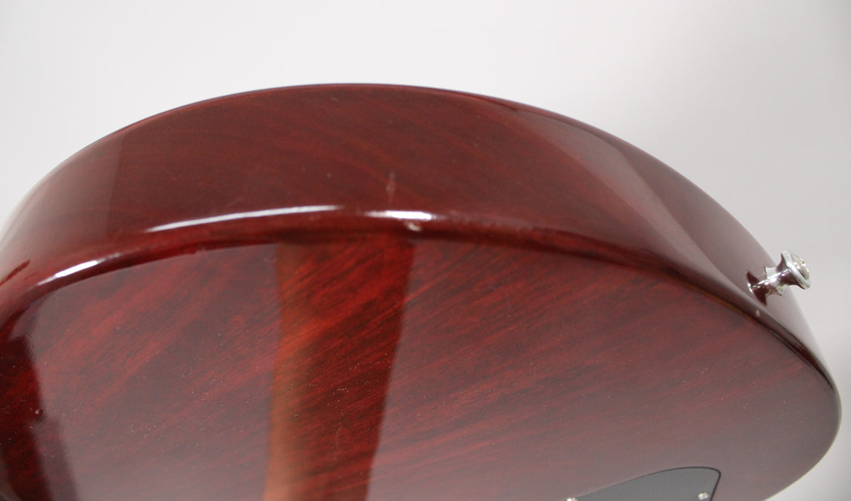 Close-up of the dark cherry guitar body's curved edge, showing a small scratch on the glossy finish and a chrome strap button.
