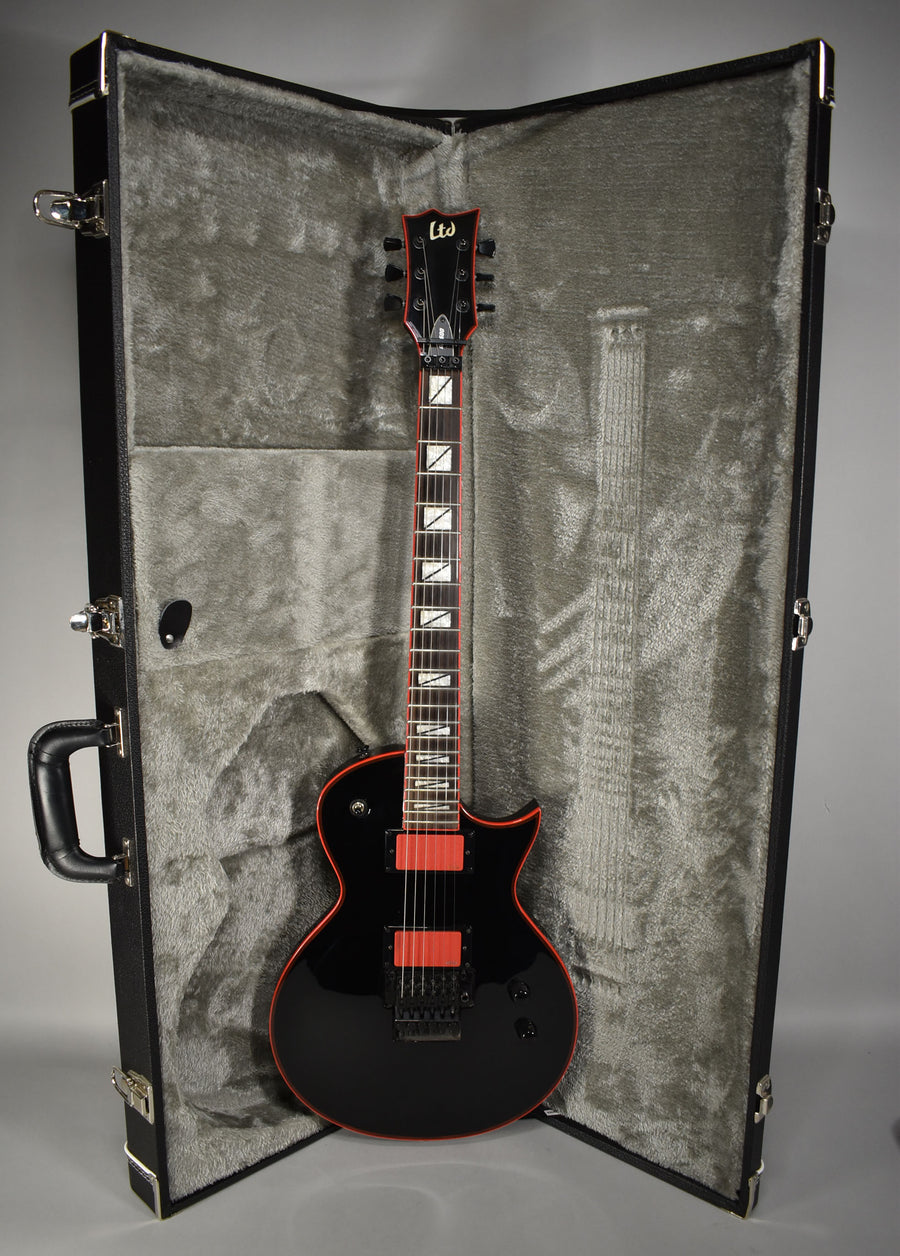 Full-length, glossy black electric guitar with red binding, red pickup covers, and a black Floyd Rose tremolo system, in a grey plush-lined case.