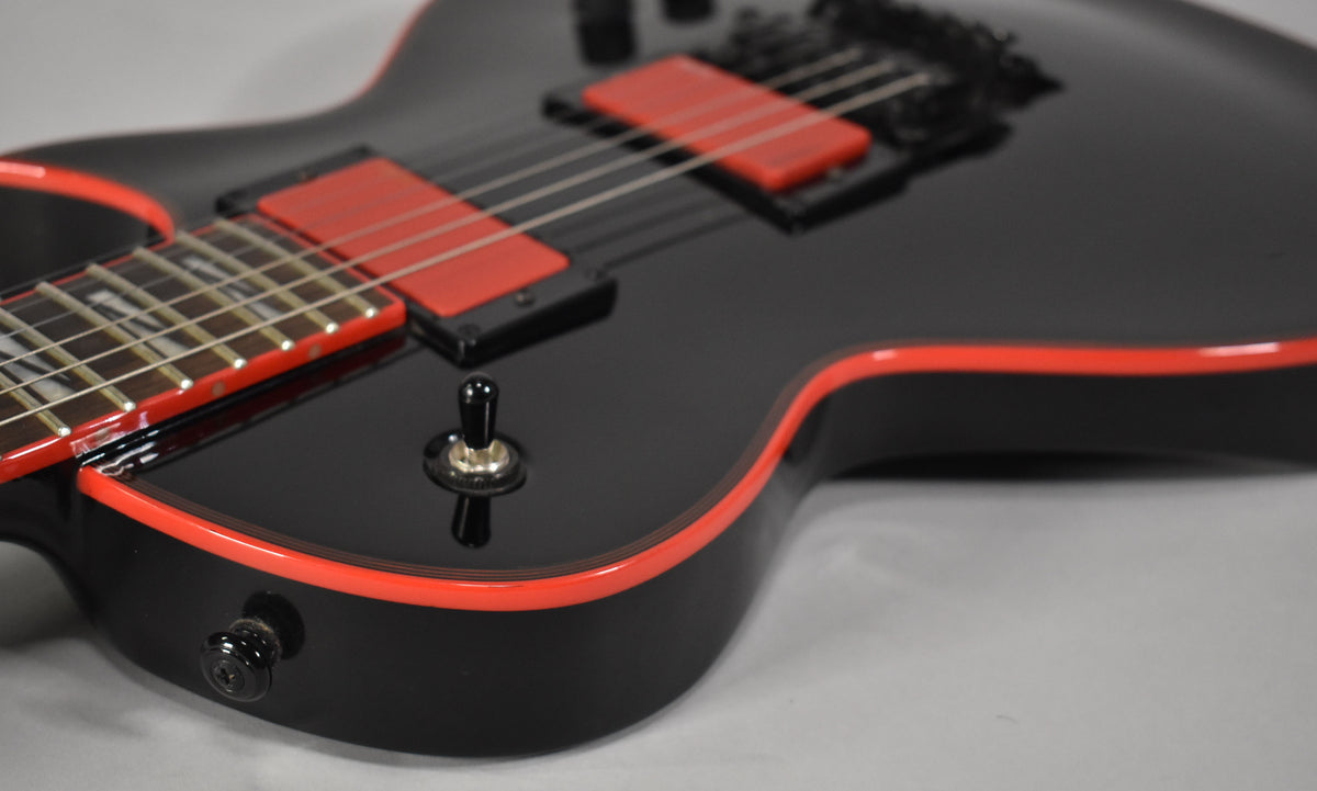 The lower body of a black electric guitar with red binding, red humbucker pickups, and a black toggle switch.