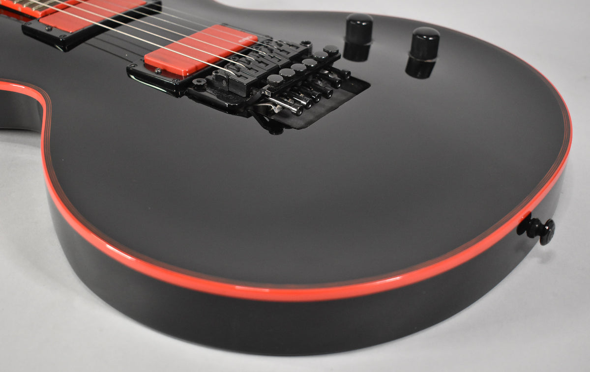 Close-up of a glossy black guitar body with red binding, showing the bridge, red humbucker pickups, and two black control knobs.