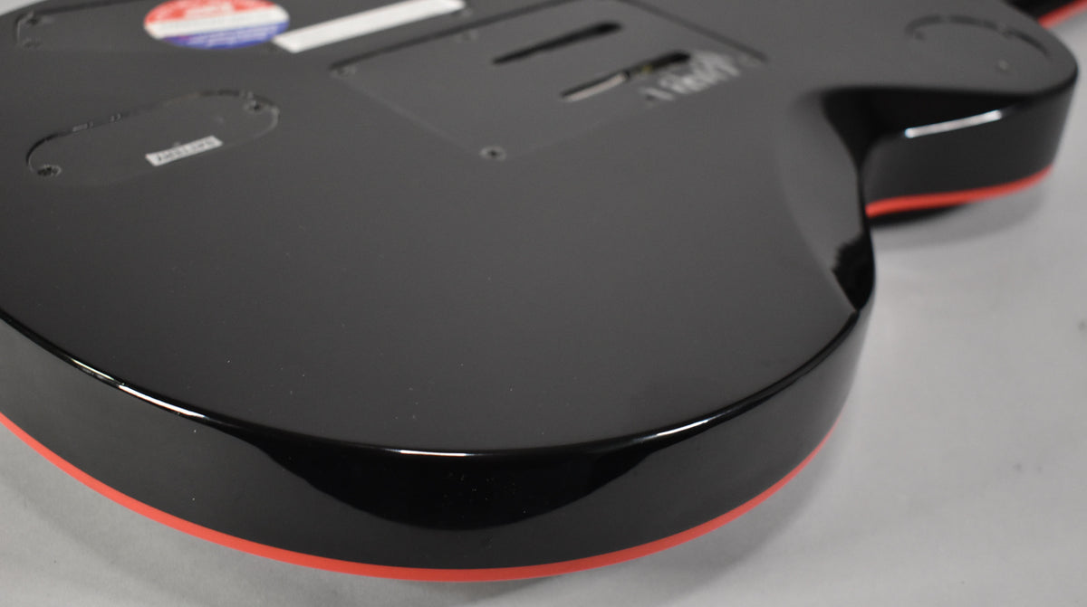 The guitar's glossy black body, with a thin red stripe along the bottom edge, is angled to show the back control plate covers.
