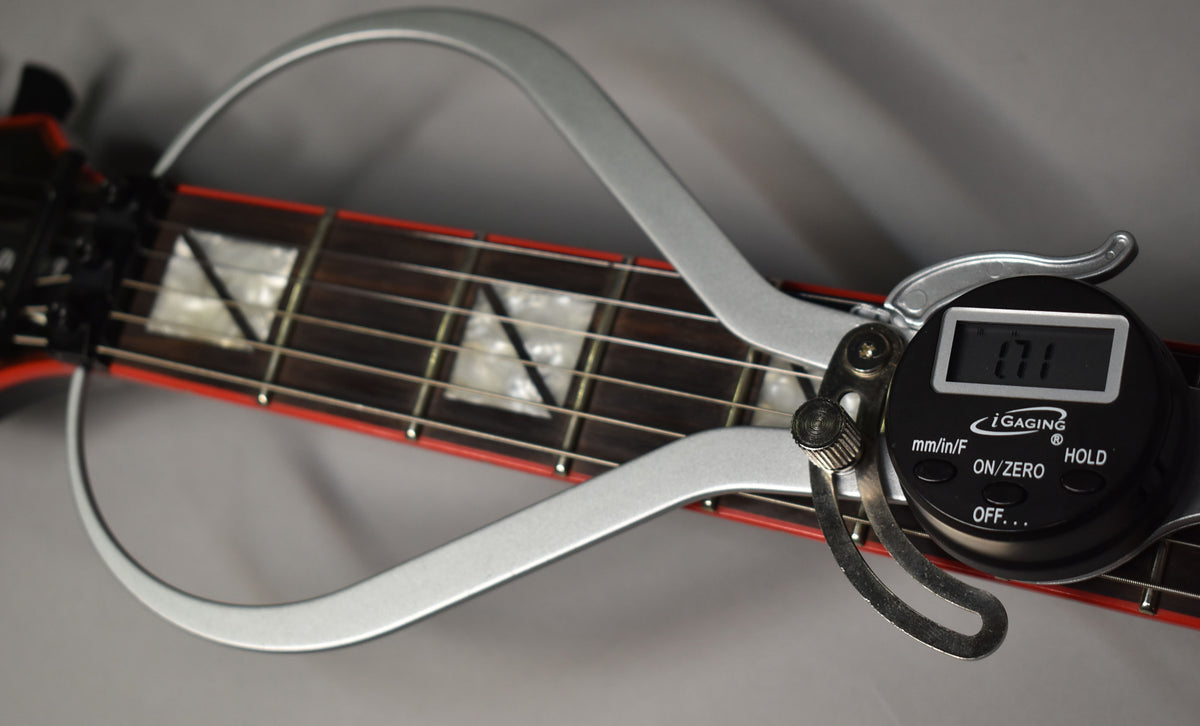 A digital caliper measures the fret height on the dark fretboard with white trapezoid inlays and red binding.