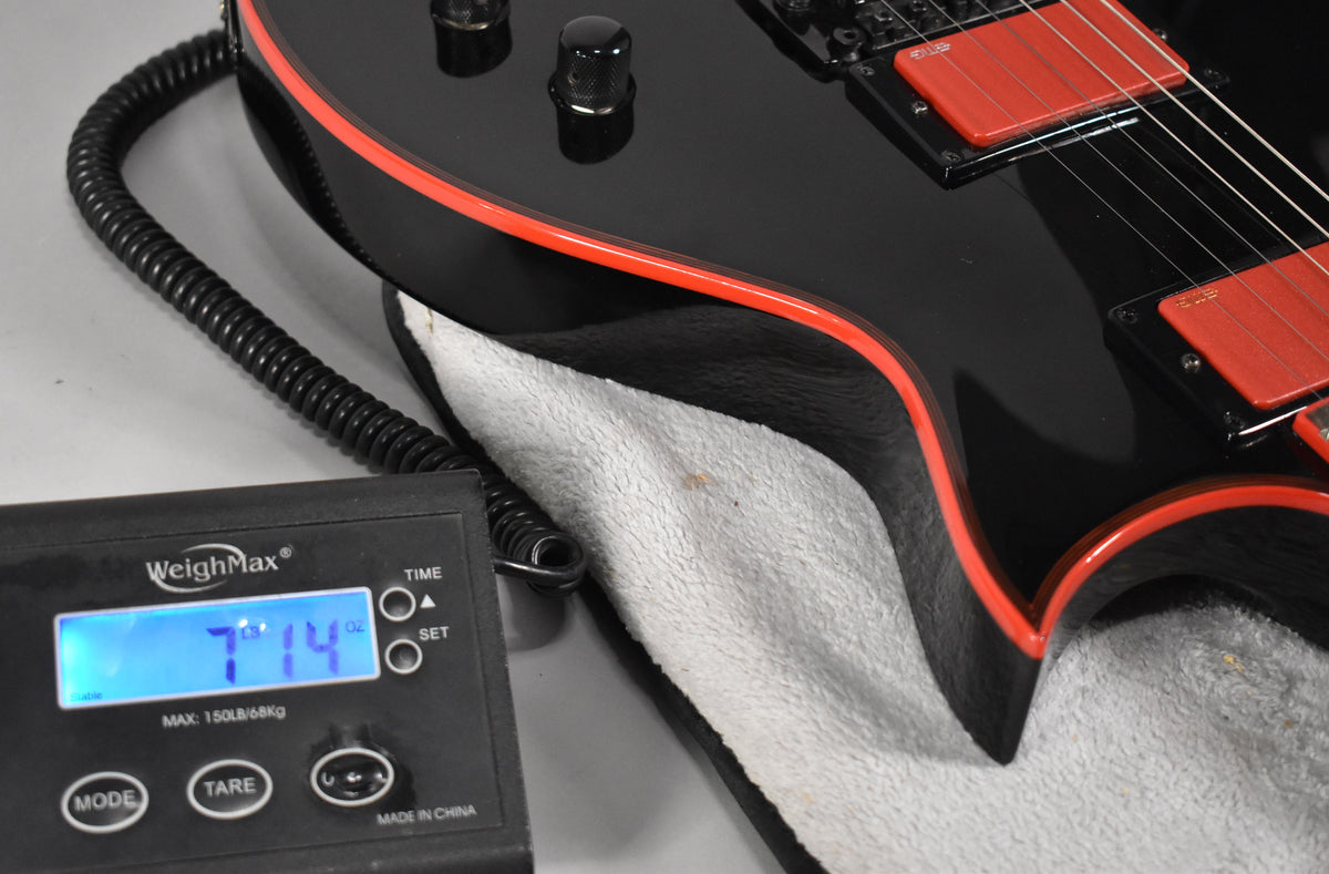 The guitar's glossy black body with red binding and red humbucker pickups is shown on a scale displaying 7 lbs 14 oz.