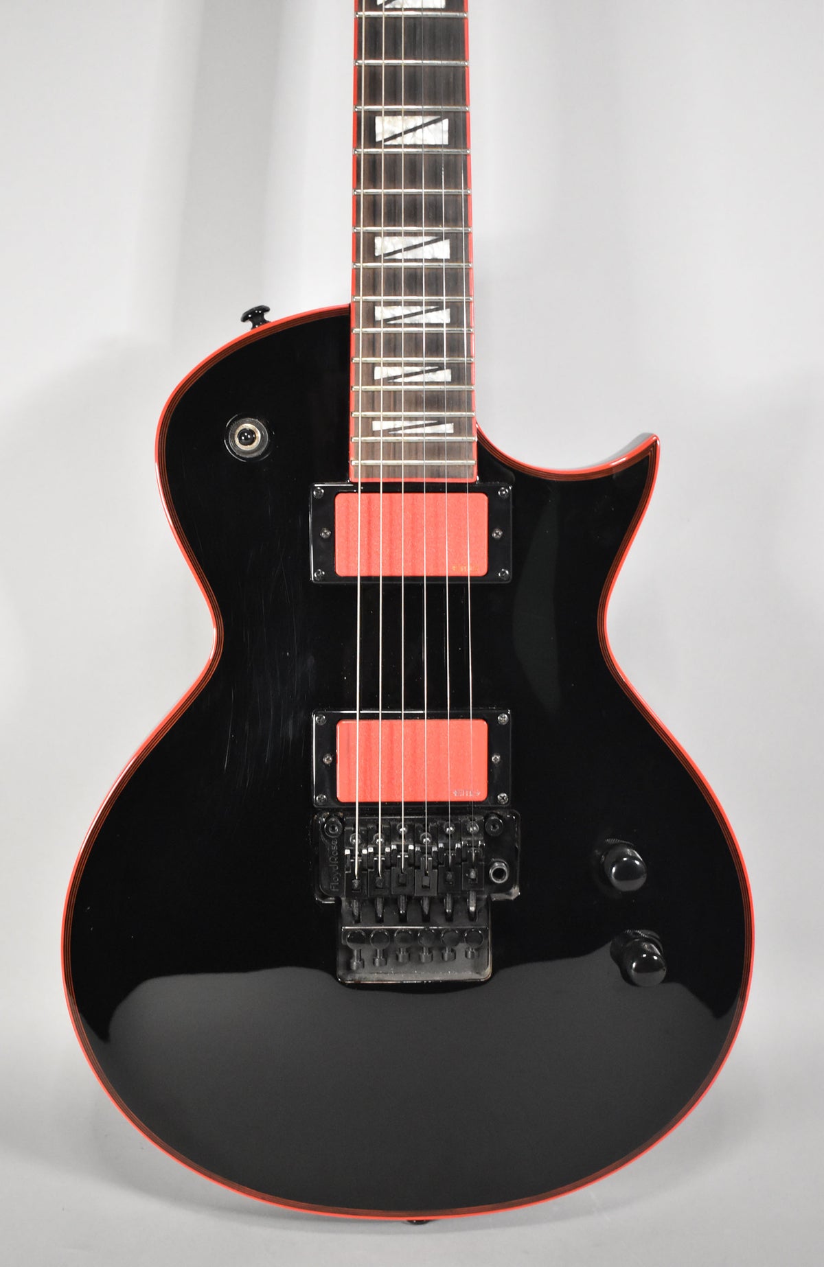 A black electric guitar with red binding and red humbucker pickups, showing a Floyd Rose tremolo system and light scratches on the body.