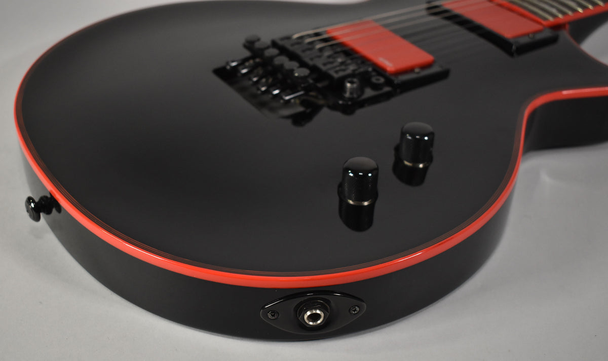 The lower bout of a black electric guitar with red binding, two black control knobs, and a black output jack.