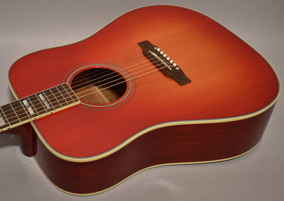 The cherry sunburst acoustic guitar's body is angled, showing the soundhole, dark wood bridge with white pins, and part of the fretboard.