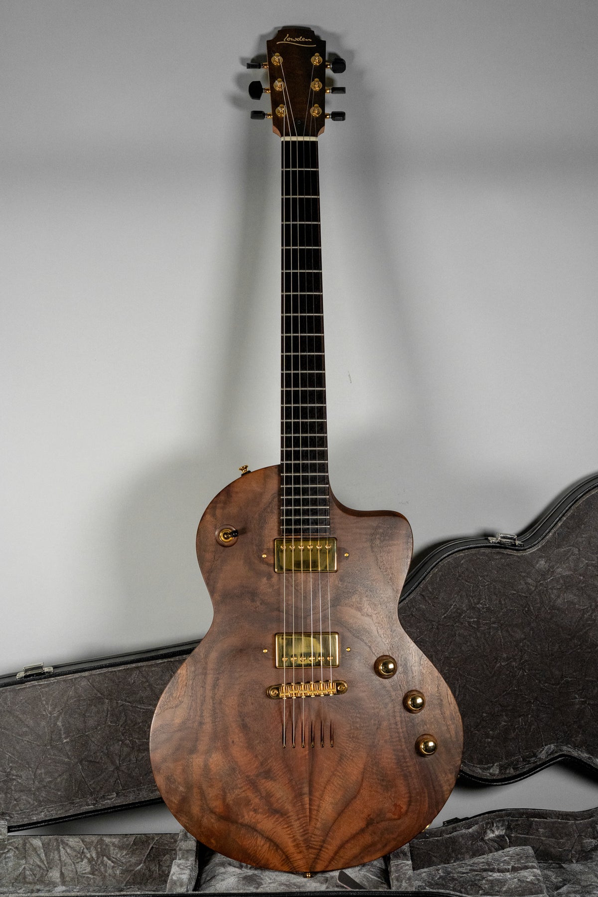 Full-length view of a dark walnut guitar with a figured grain, gold hardware, and three control knobs, standing in its open case.
