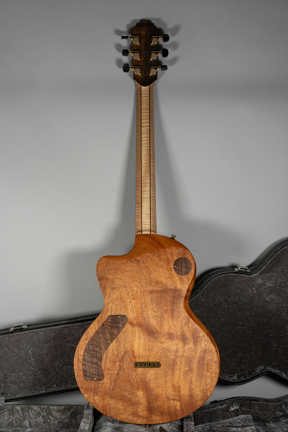 The back of a light brown wooden guitar with a figured walnut control plate and a natural maple neck, gold tuners.