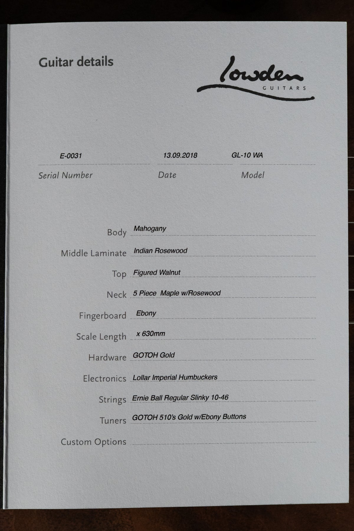 Close-up of a white paper spec sheet with black text, detailing guitar features like "Figured Walnut" top and "GOTOH Gold" hardware.