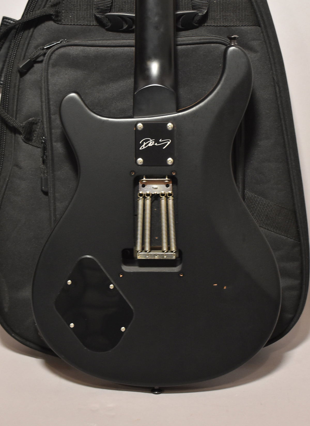Rear view of a matte black electric guitar, showing the neck joint, tremolo springs, control cavity cover, and minor finish wear.