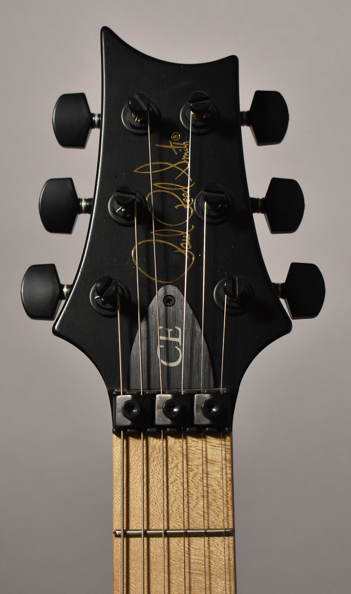 Close-up of the guitar's matte black headstock with gold script, black tuners, and a locking nut over a light wood fretboard.