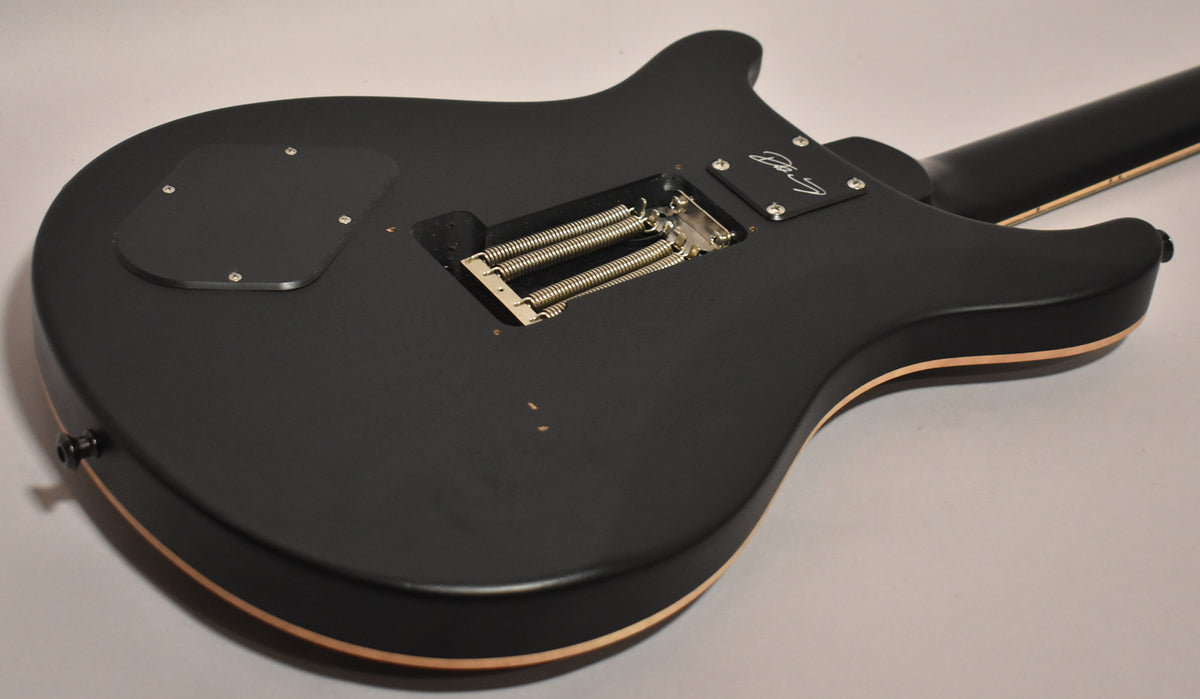 The back of a matte black electric guitar, angled to show the tremolo spring cavity and a few small dings.