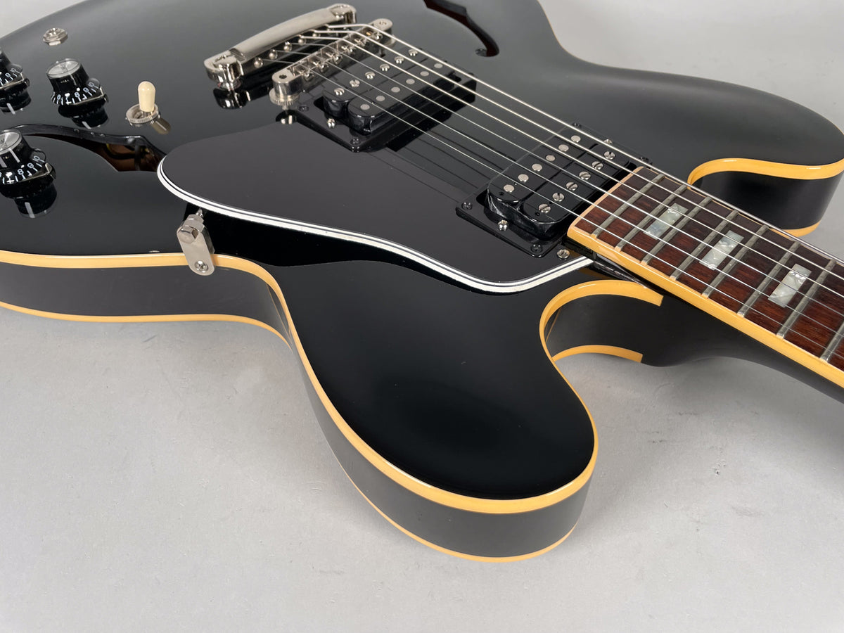 Close-up of a glossy black hollow-body electric guitar, showing the lower bout, pickguard, bridge, neck, and controls.