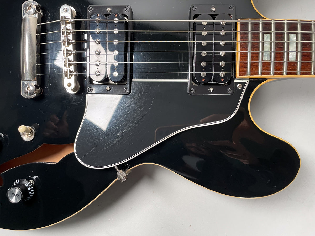Close-up of a black semi-hollowbody guitar's lower bout, showing the bridge, two humbuckers, pickguard scratches, and a volume knob.