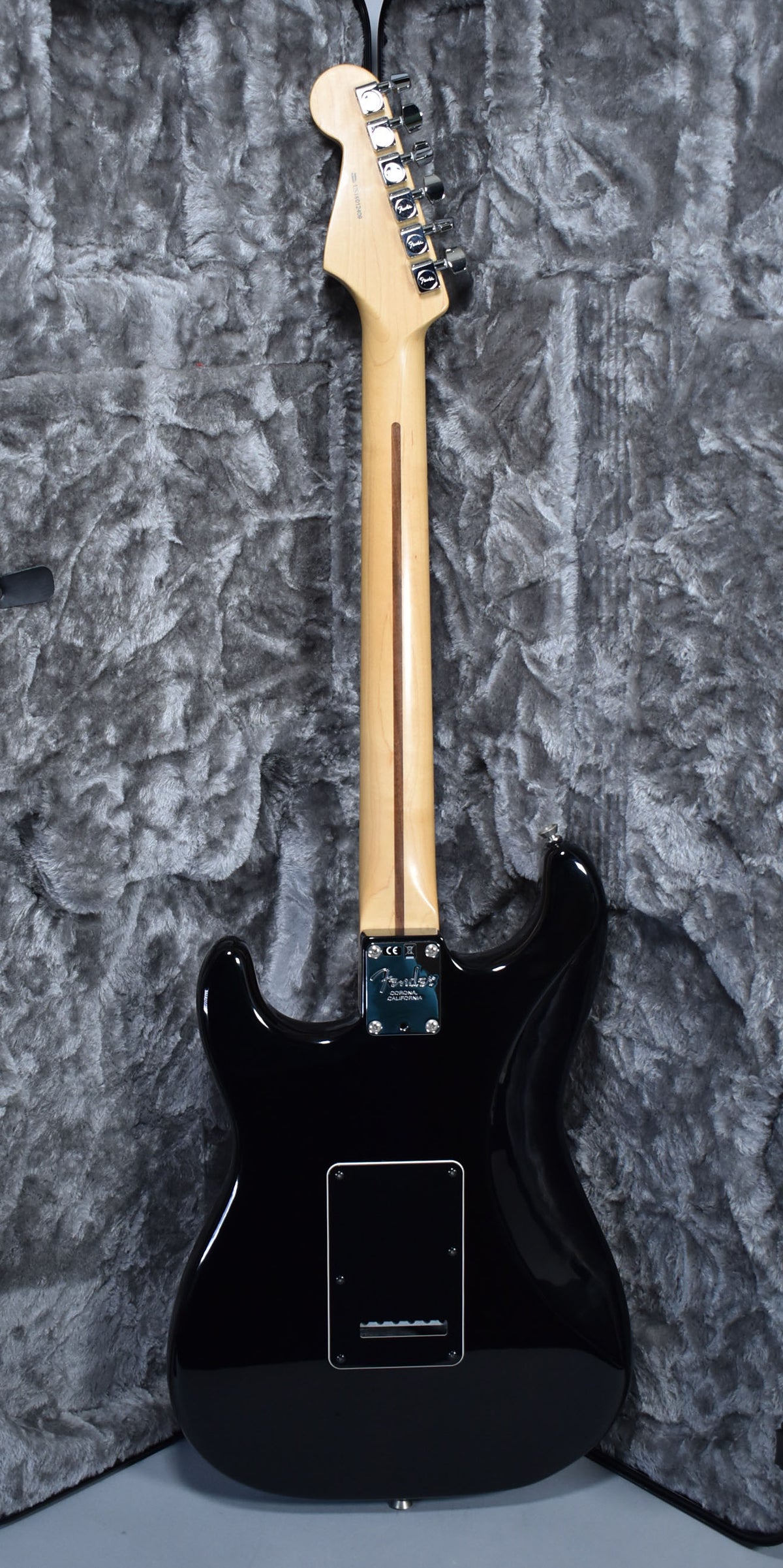 The back of a glossy black Stratocaster with a light maple neck, chrome tuners, and a black tremolo cover, resting in a grey plush case.