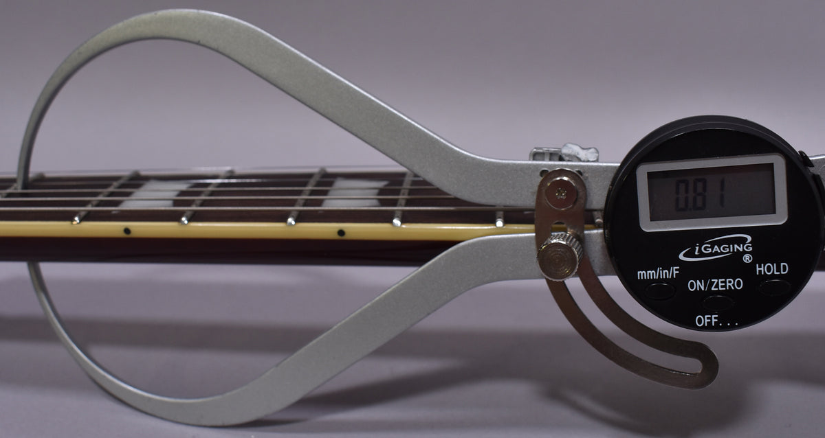 A digital caliper measures the guitar's fretboard, showing 0.81mm, with block inlays and cream binding visible on the dark wood neck.