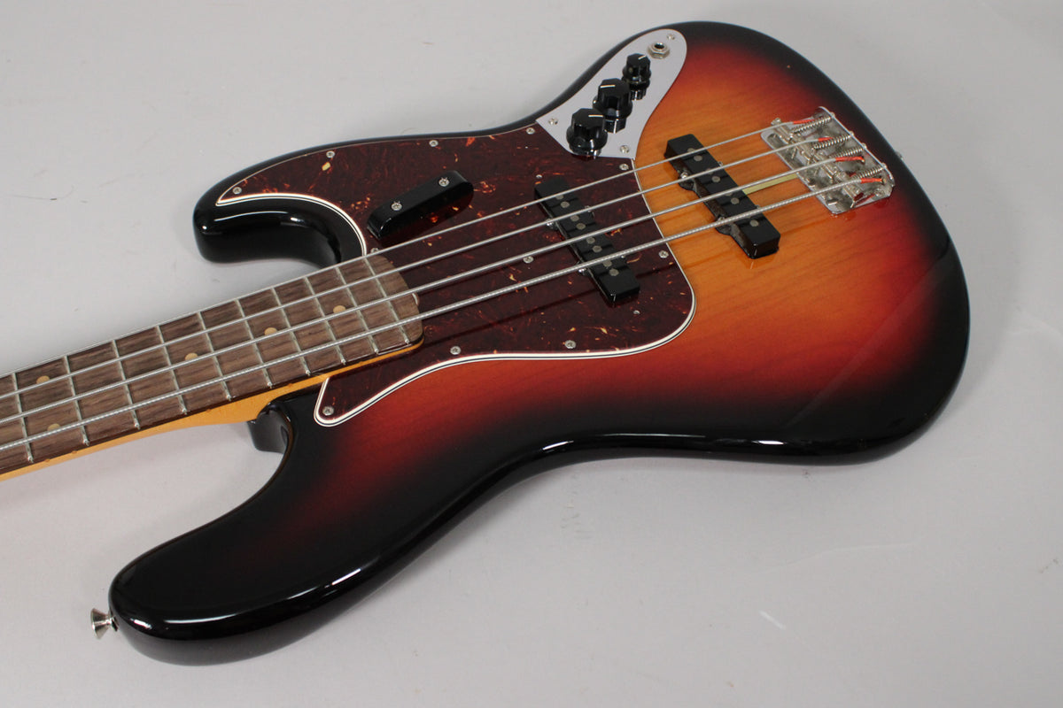 The 3-color sunburst Jazz Bass is angled from the lower left, showing the glossy body, tortoise pickguard, and chrome bridge.