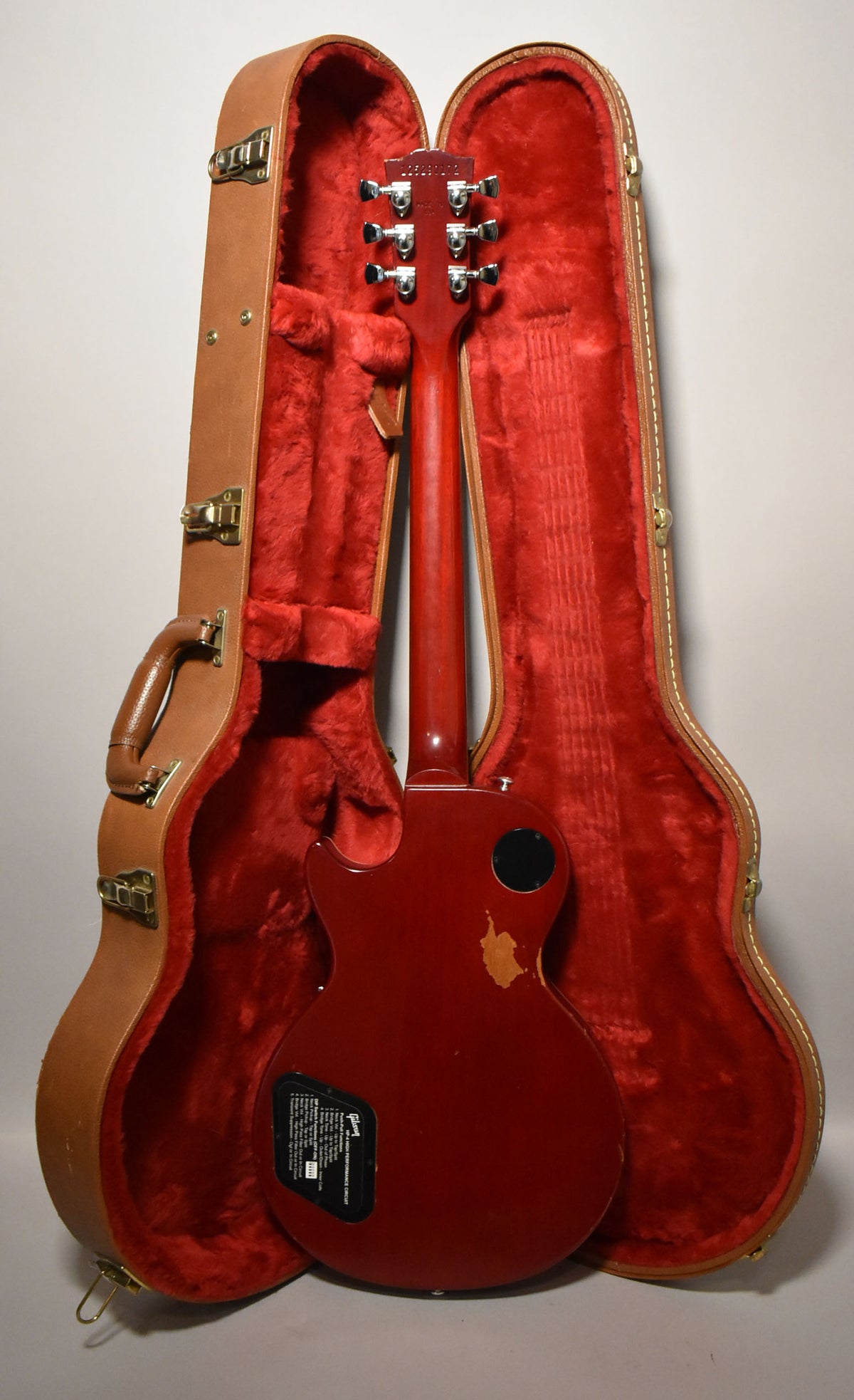 The wine red Les Paul is shown from the back, resting in its open tan hard case with red plush lining, showing wear on the body.