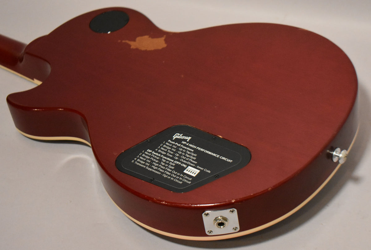 The back of a wine red guitar shows a black control cavity cover with white text, a strap button, and input jack.