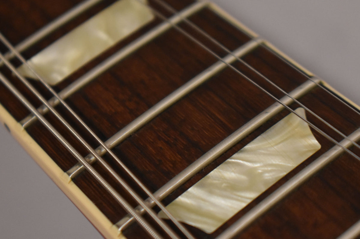Close-up of the dark rosewood fretboard with pearloid trapezoid inlays, showing the frets and strings at an angle.
