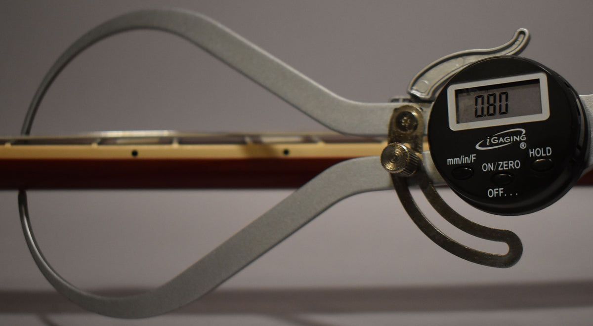 A digital caliper measures the guitar's neck, showing 0.00 on its display, with the dark wine red neck and light fretboard visible.