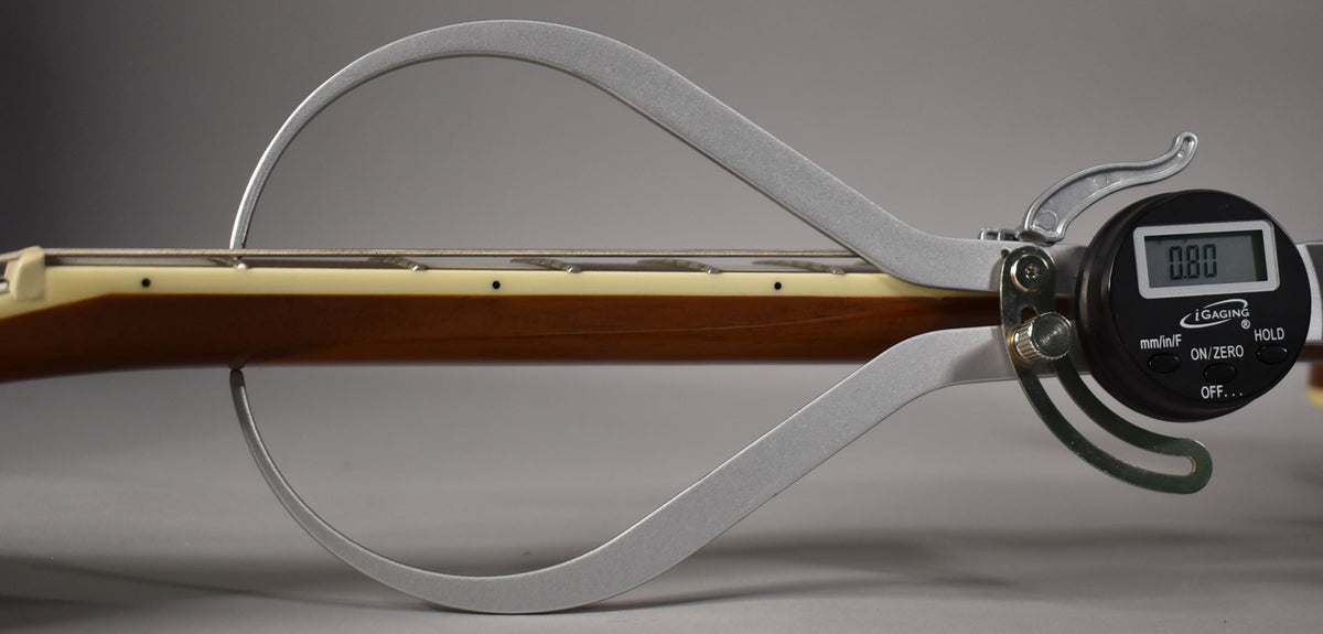 A digital caliper measures the guitar's neck relief, showing a brown neck, cream binding, and dark fretboard with dot inlays.