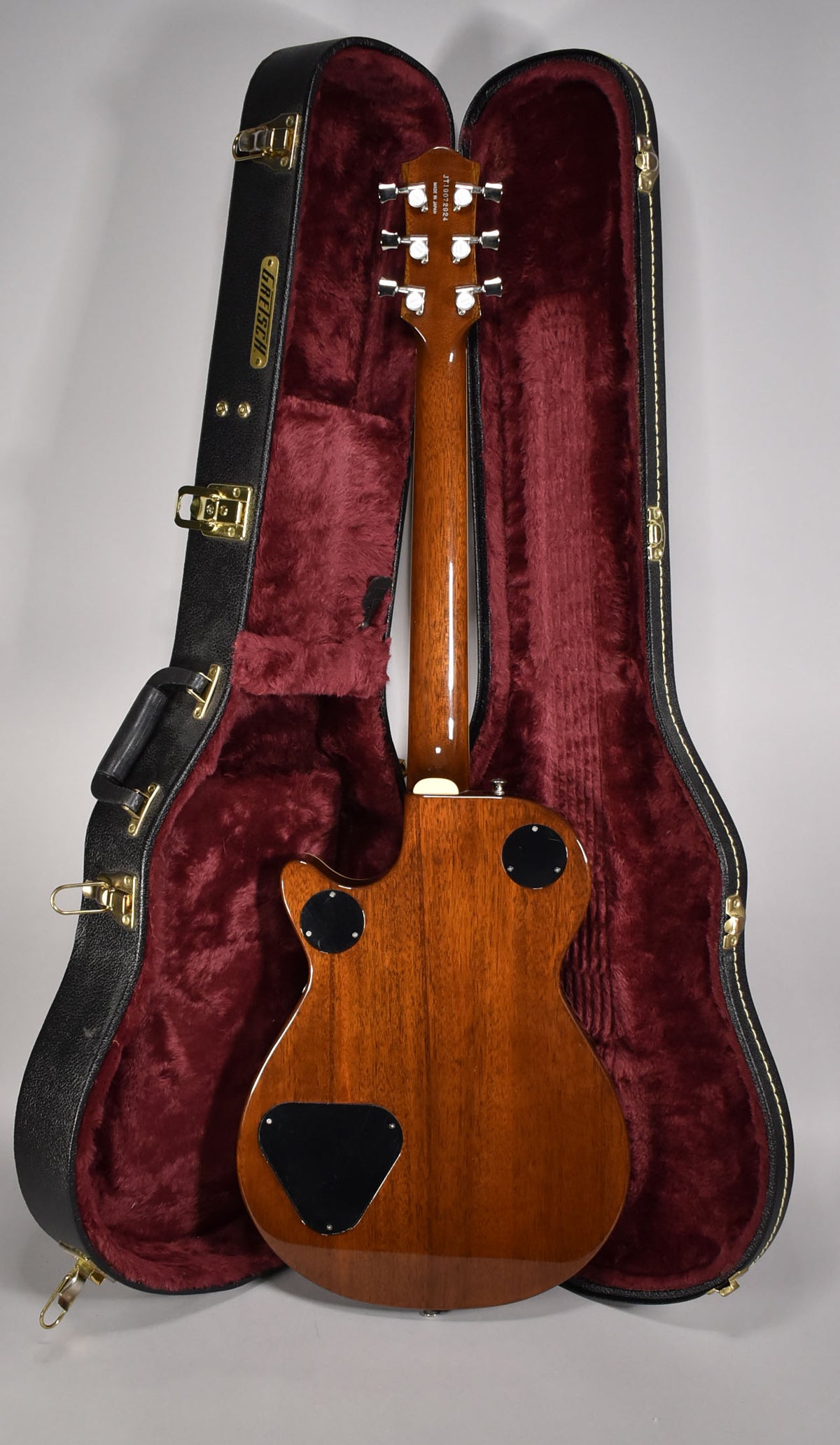 The guitar's glossy brown back and neck are visible, with three black control plates, resting in a black hard case with red plush lining.