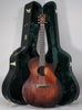 A dark brown acoustic guitar with a cutaway body stands upright in front of its open, dark green-lined hard case.