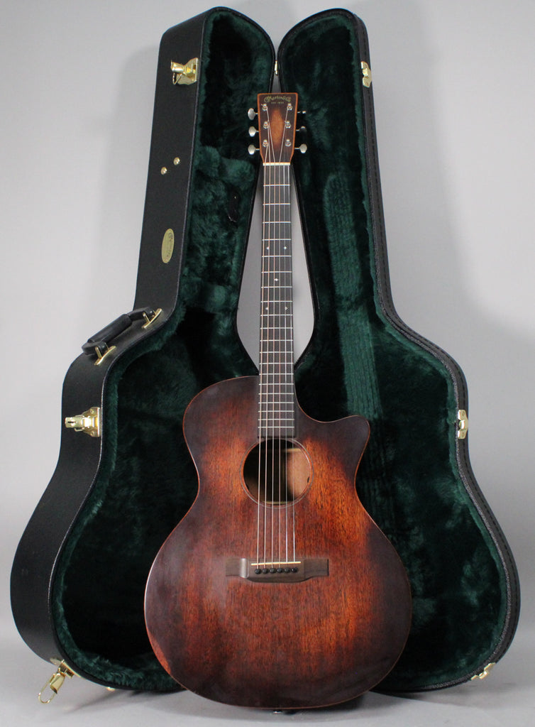 A dark brown acoustic guitar with a cutaway body stands upright in front of its open, dark green-lined hard case.