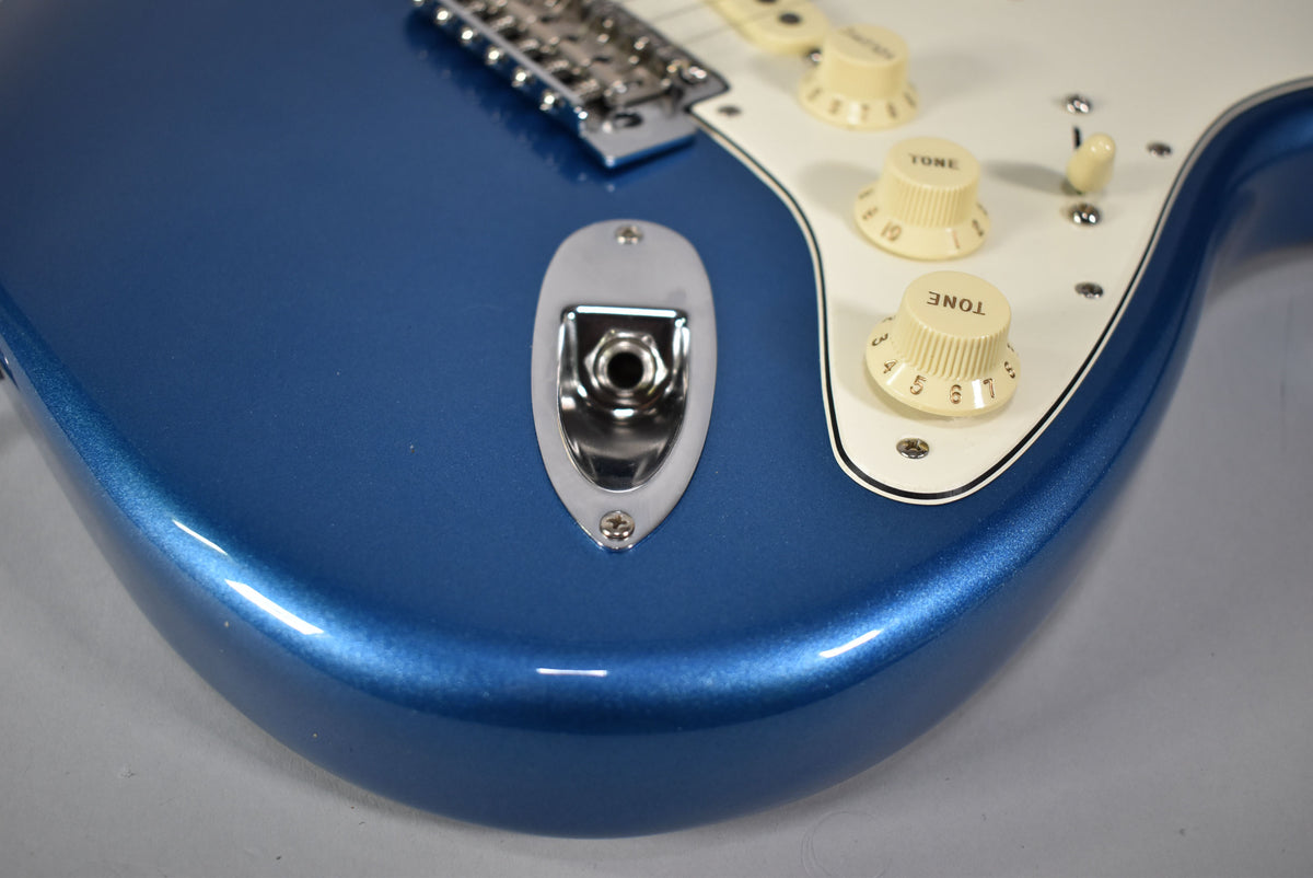 Close-up of a blue guitar body showing the input jack, part of the bridge, and two cream-colored tone knobs on a white pickguard.