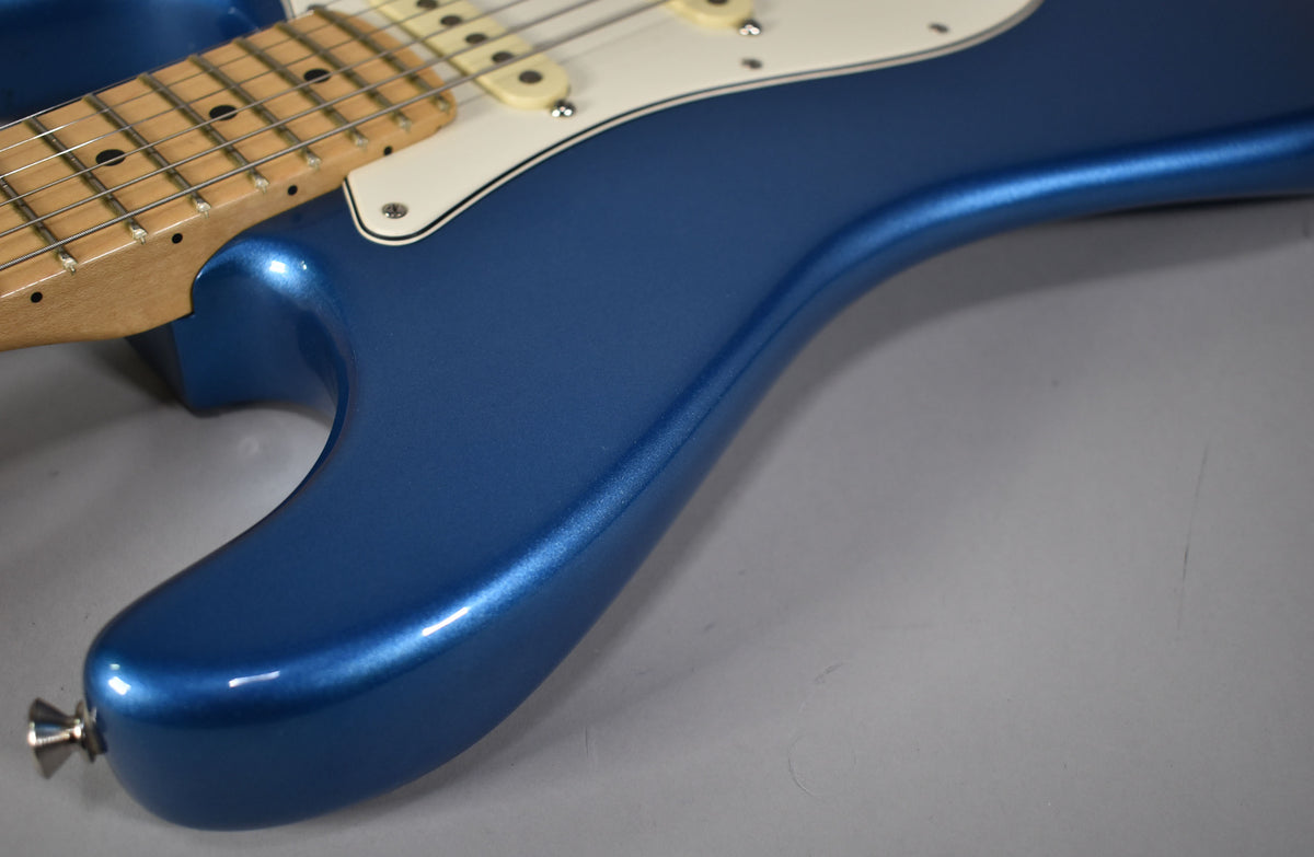 The lower horn and body of a metallic blue Stratocaster are shown, with a maple neck and white pickguard visible at the top left.