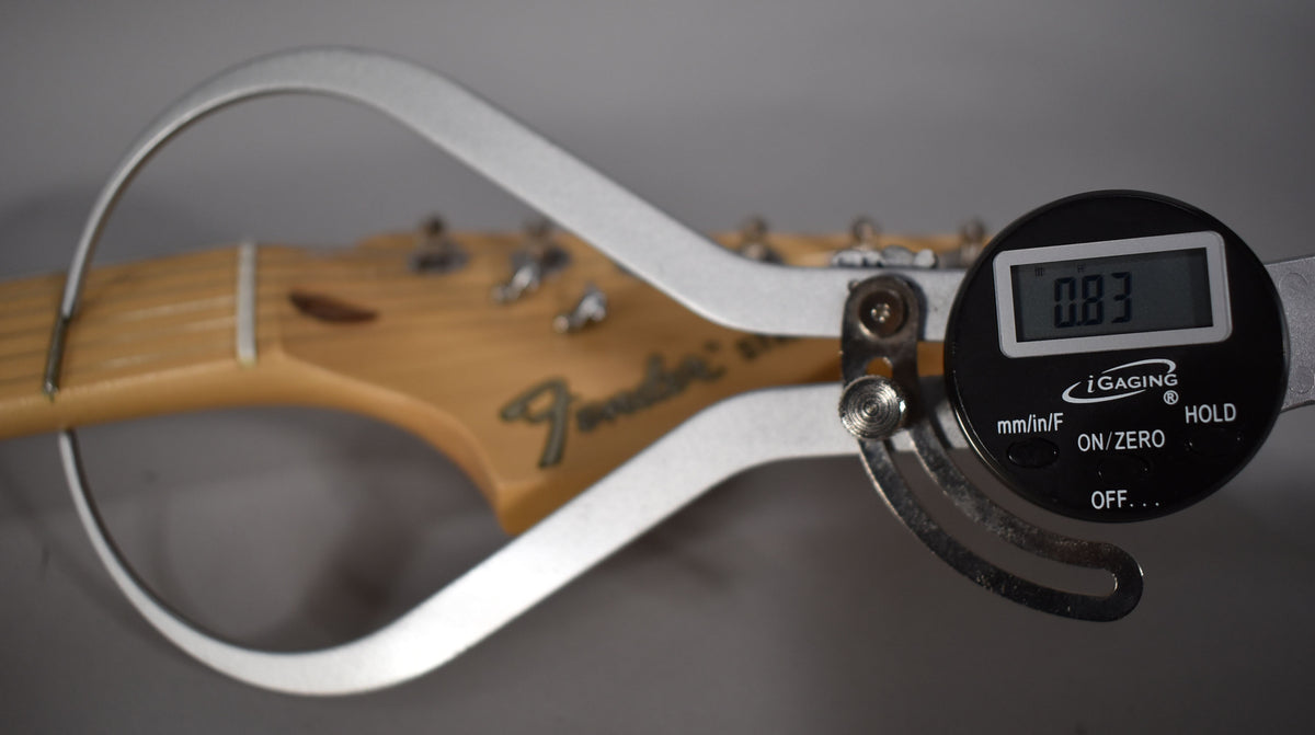 A digital caliper measures the guitar's natural wood headstock, showing 0.83 on its screen, with tuning pegs visible.