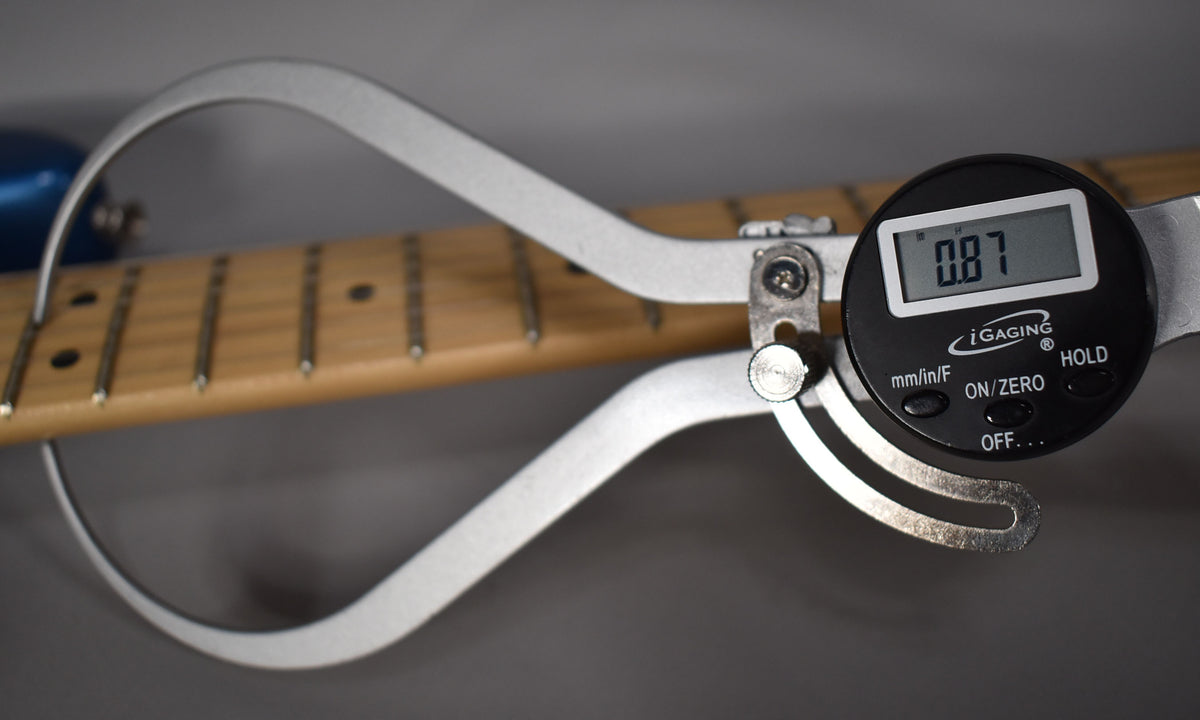 A digital caliper measures the maple fretboard's thickness at 0.87 inches, with a blue guitar body visible in the background.
