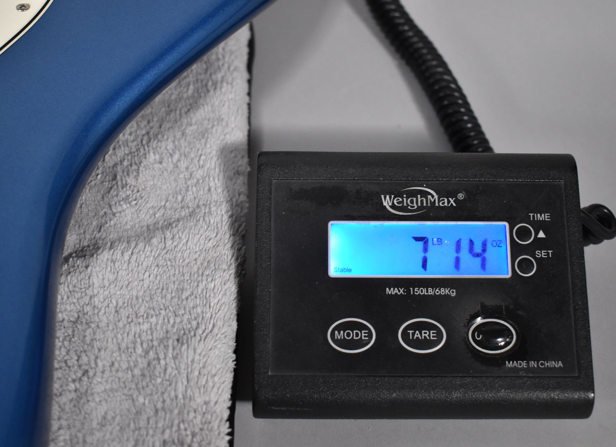 The guitar's satin Lake Placid Blue body rests on a towel next to a scale displaying its weight: 7 lbs, 14 oz.