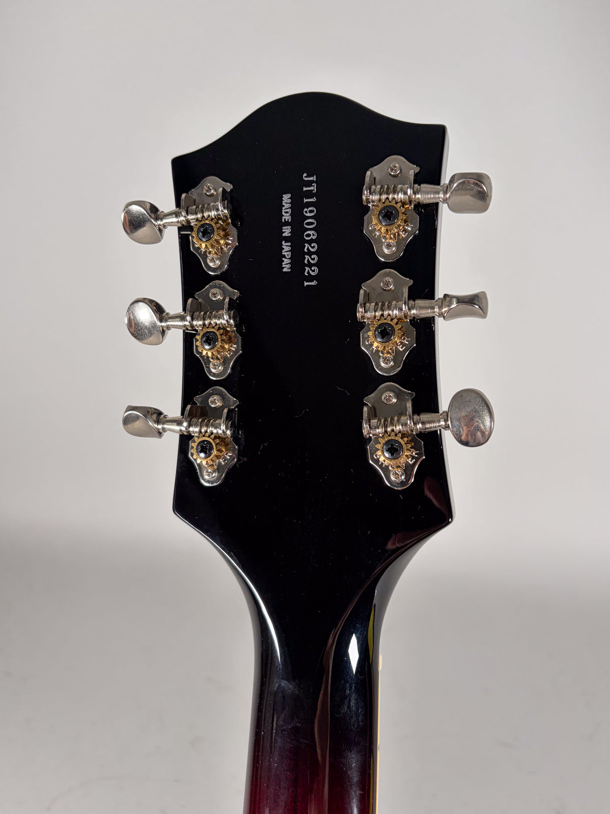 The guitar's glossy black headstock, with six chrome tuners featuring gold gears, is viewed from the rear.