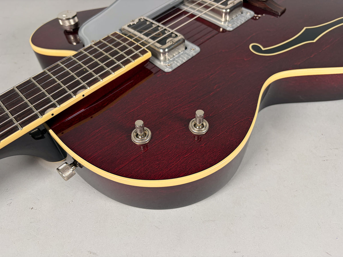 Close-up of the deep red hollow body, showing two chrome toggle switches, a bound f-hole, and the lower fretboard.