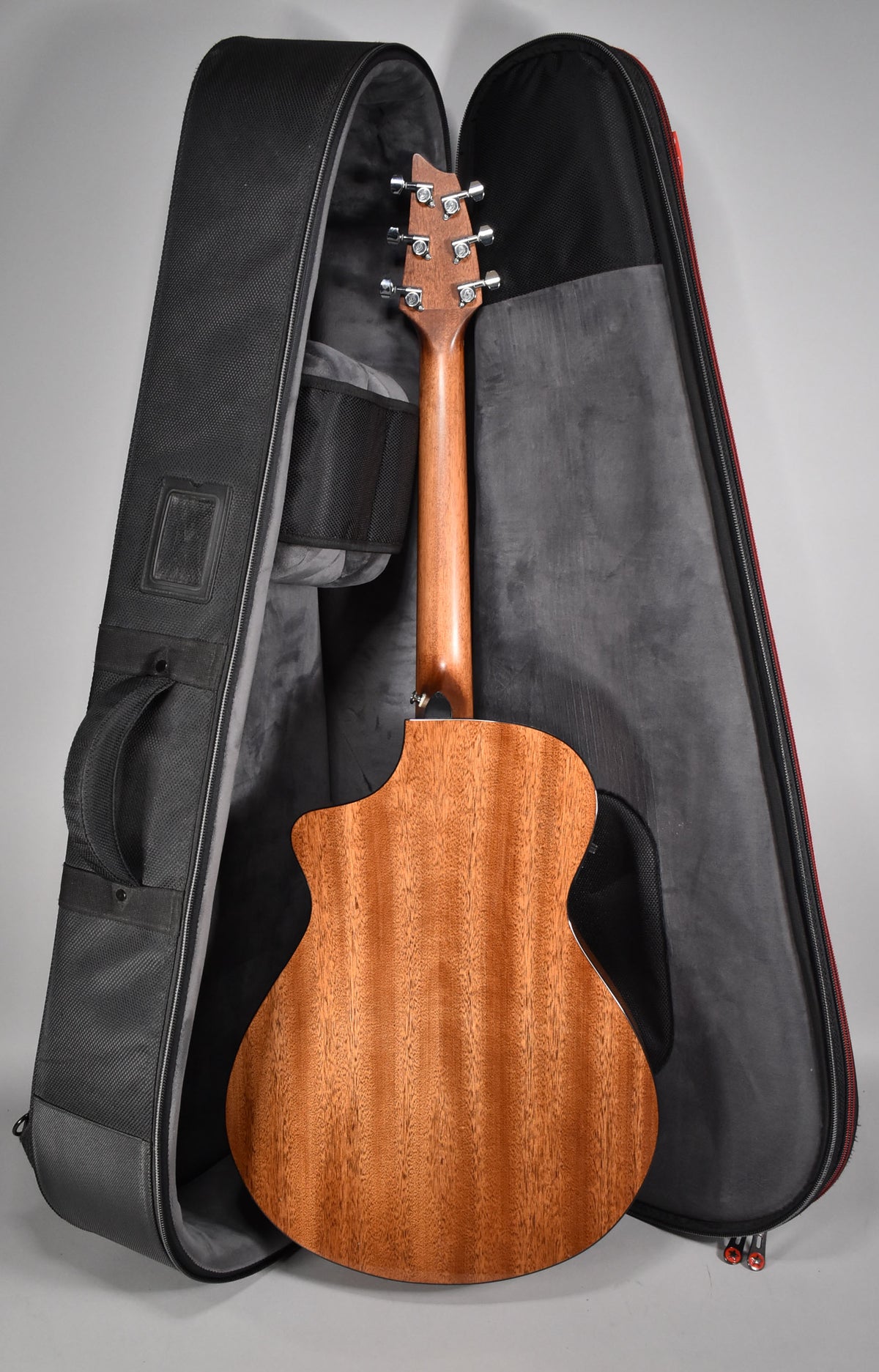 The acoustic guitar's mahogany back and neck are visible, with six chrome tuners on the headstock, standing in an open black gig bag.