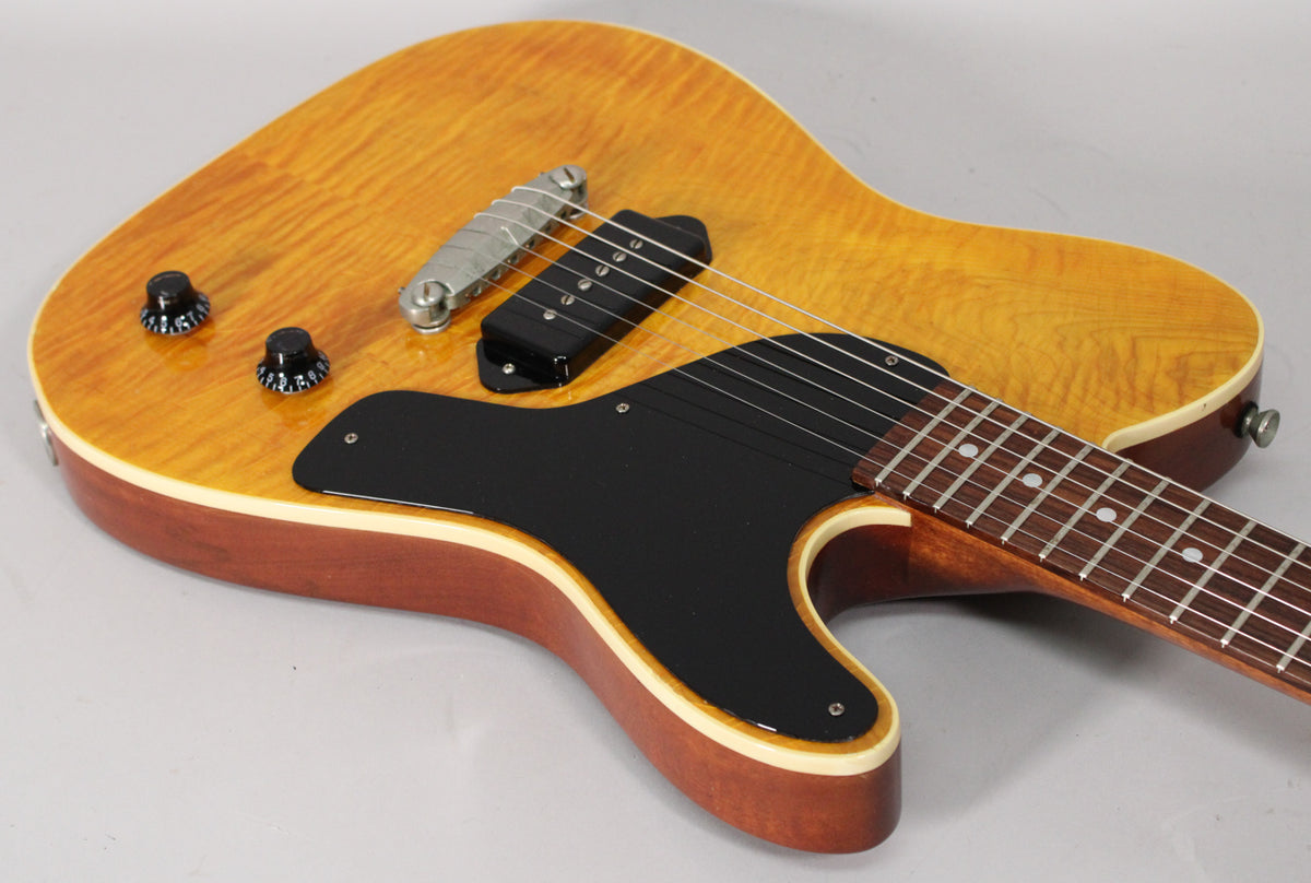The amber nitro guitar body, with a black pickguard and P90 pickup, is angled from the lower left, showing two black control knobs.