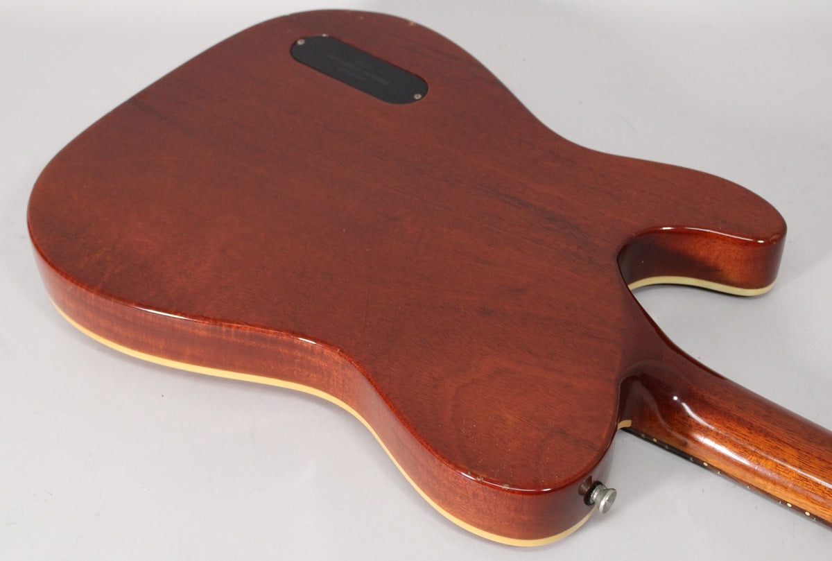 The back of an amber nitro guitar body with cream binding, showing a black control cavity cover and a chrome strap button.