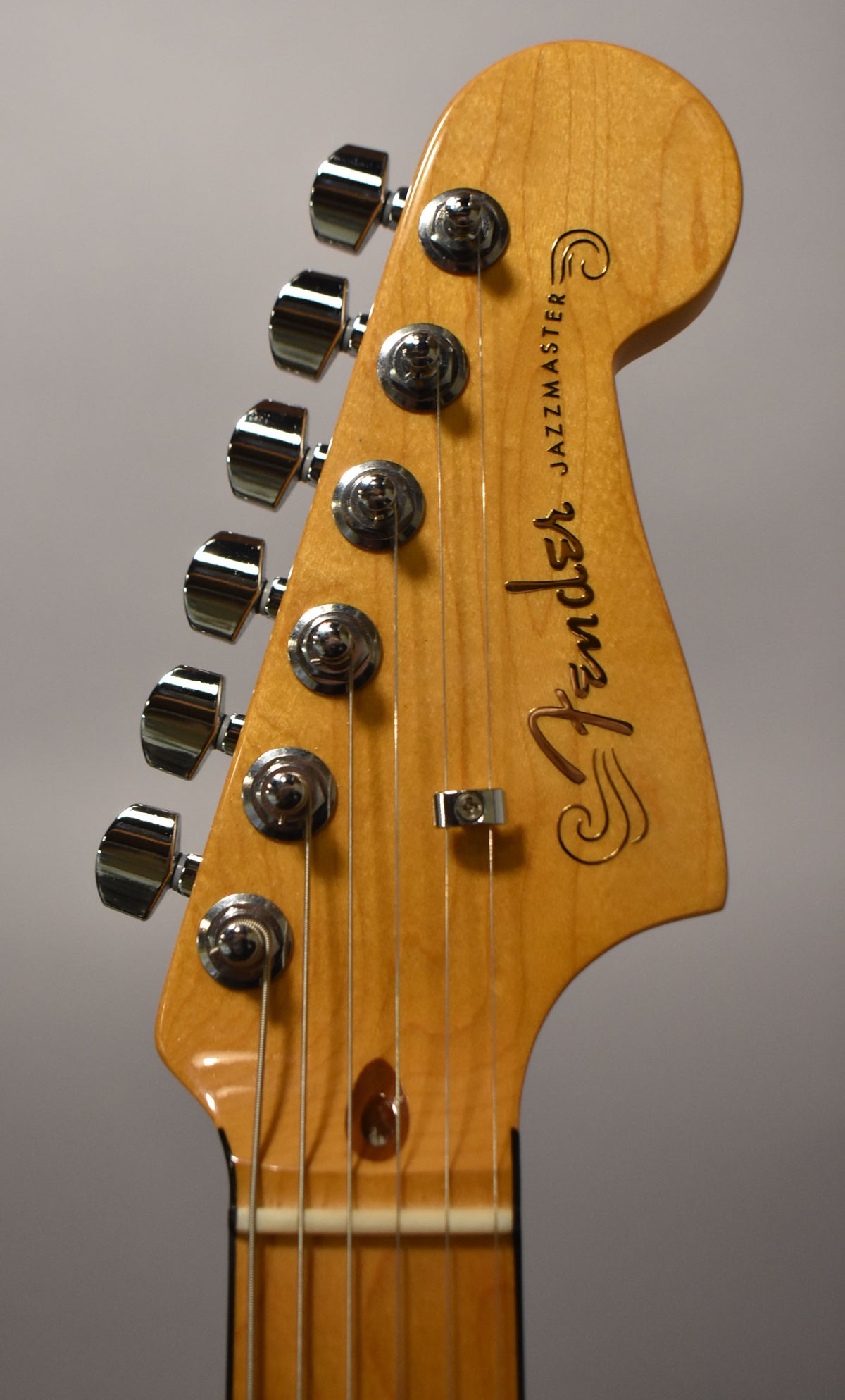 Close-up of the Jazzmaster's light maple headstock with a glossy finish, showing six chrome tuners and a string tree.