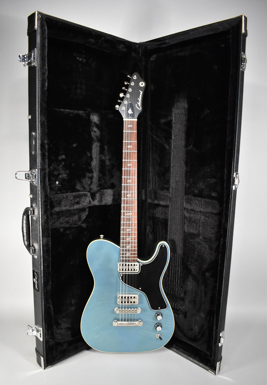Full view of a metallic Pelham blue guitar with a black pickguard and aged hardware, standing in its open, black-lined hard case.