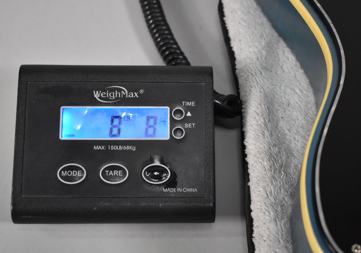 A WeighMax scale displays "8 LB 8 OZ" with a Pelham Blue guitar body, its cream binding, and a soft grey cloth visible.