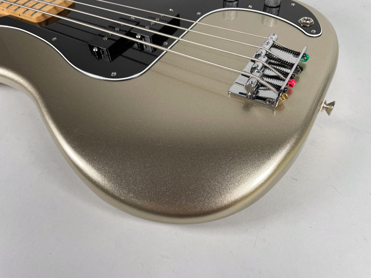 Close-up of a metallic gold bass guitar body with a black pickguard, chrome bridge, and four strings with colored ends.