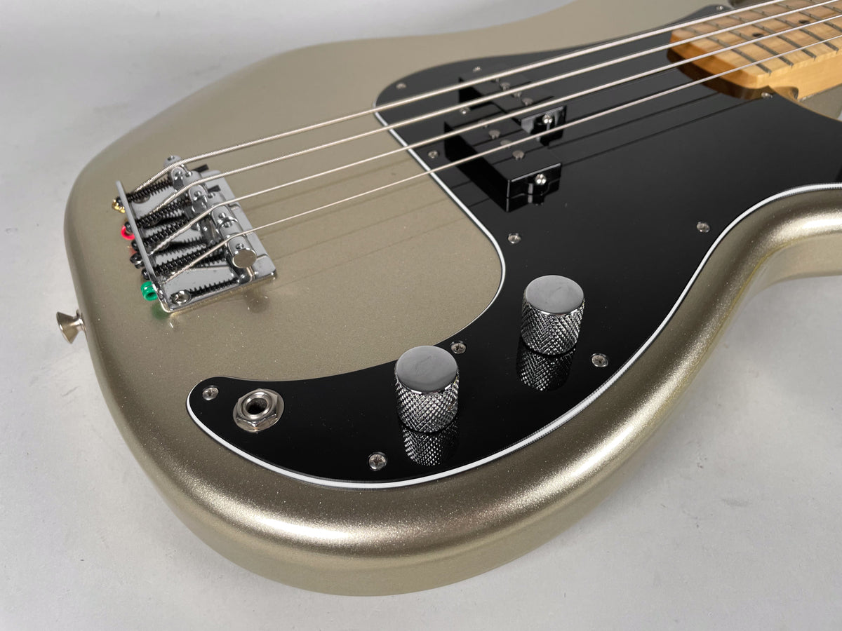 Close-up of a metallic gold bass guitar body with a black pickguard, chrome bridge, two knurled chrome knobs, and an input jack.