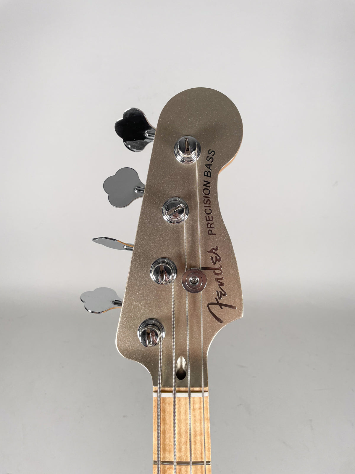 Close-up, angled view of the bass headstock in metallic champagne with four chrome clover-leaf tuners and a light maple neck.