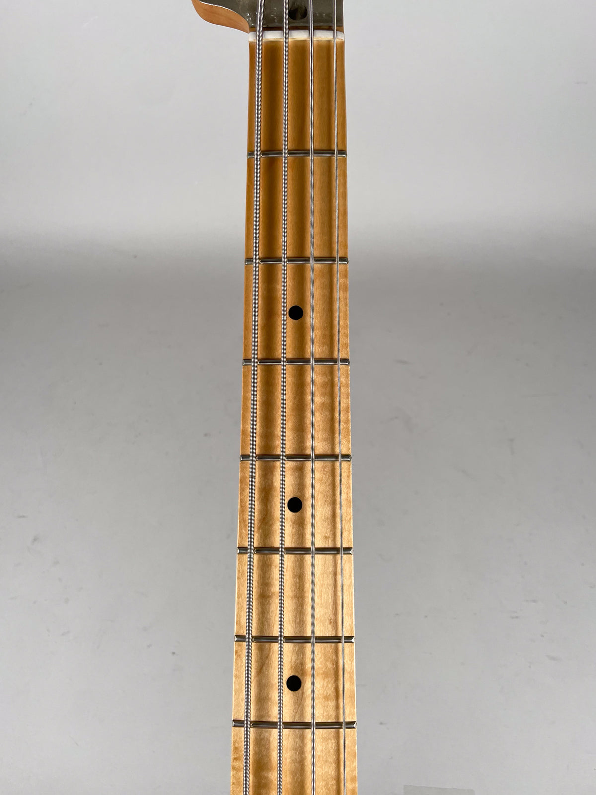A close-up, straight-on view of the bass guitar's maple fretboard, showing black dot inlays and silver frets with four strings.