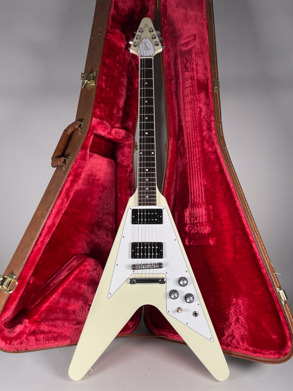 A white V-shaped electric guitar with a white pickguard and black humbuckers, standing in a brown hard case with red plush lining.