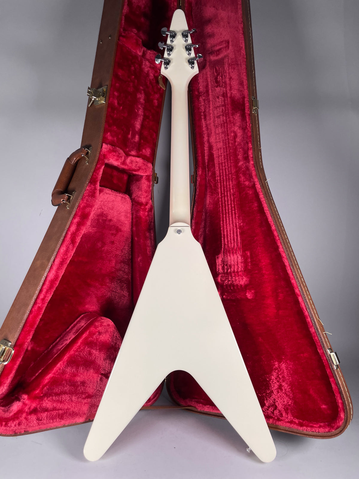 The back of the white Flying V guitar is shown standing upright in its open brown case with red plush lining, revealing six chrome tuners.