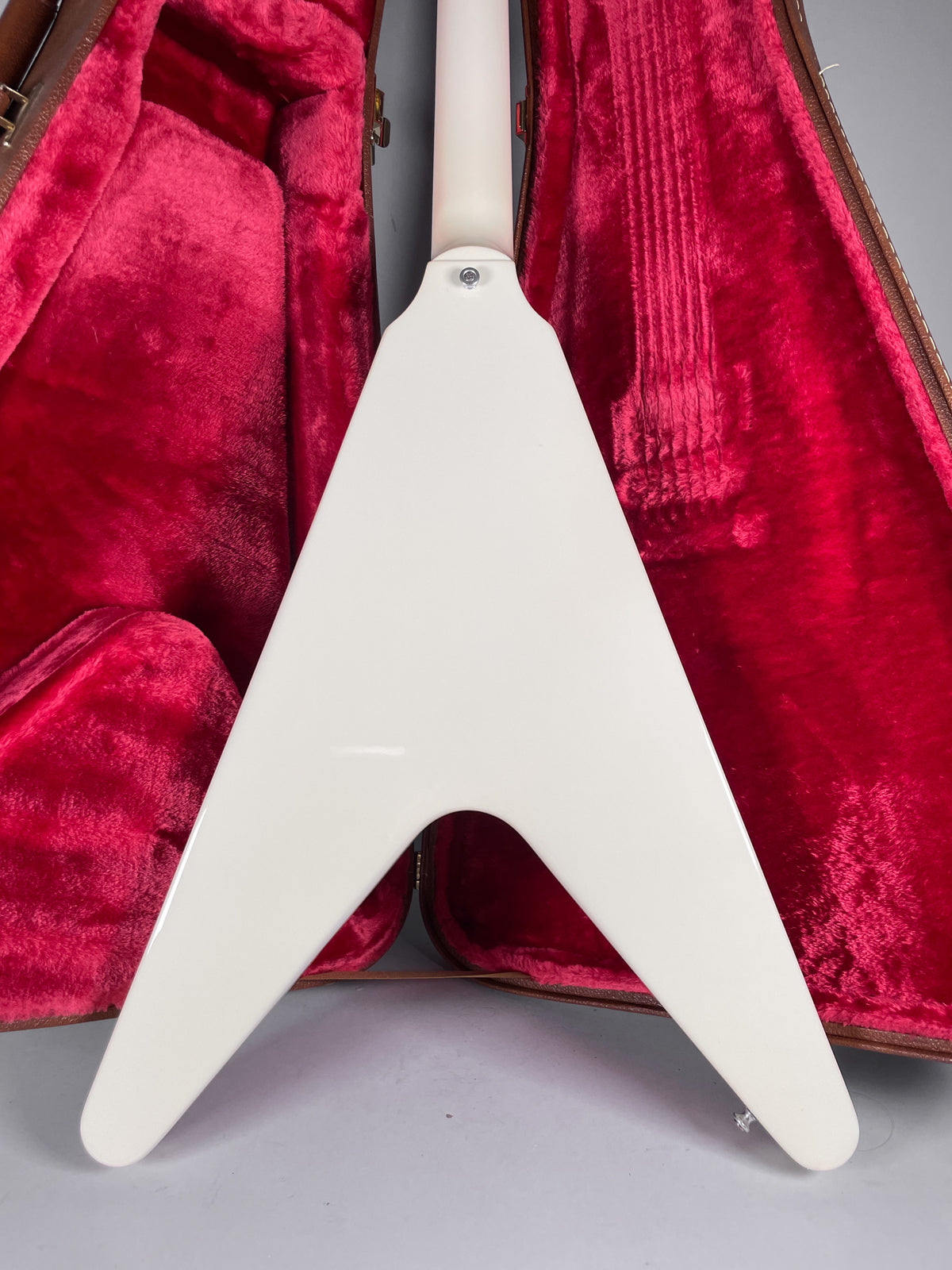 The back of a white Flying V guitar, showing the neck joint and strap button, resting in a red velvet-lined case.