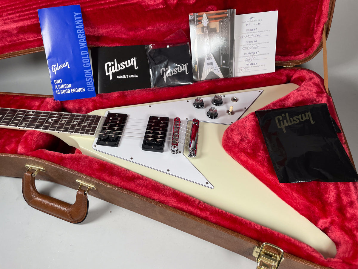 The white Flying V guitar rests in its red-lined brown hard case, with manuals and paperwork visible above the body.